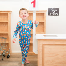 Load image into Gallery viewer, Child in colorful pajamas standing in a kitchen with wooden cabinets and a sign in the background.