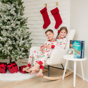 Two children in matching pajamas sitting on a chair next to a Christmas tree and stockings.